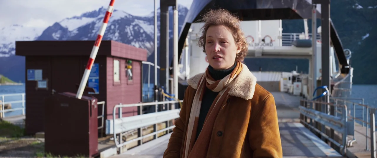 More Than Ever (2022) movie still: medium — A metal ferry landing ramp sits before a large vessel in a Norwegian fjord. A woman in a brown winter…
