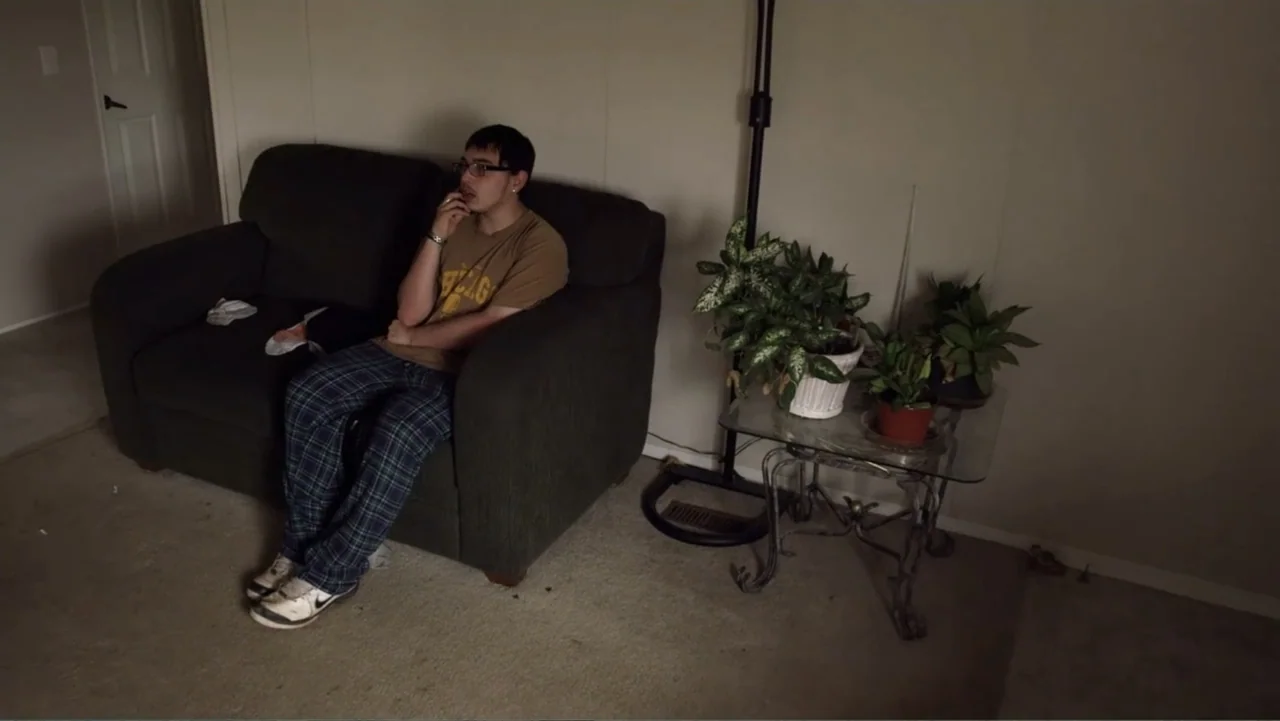 Rich Hill (2014) movie still: medium — A sparsely furnished living room features a dark sofa, a glass side table, and plants. A man sits on the…