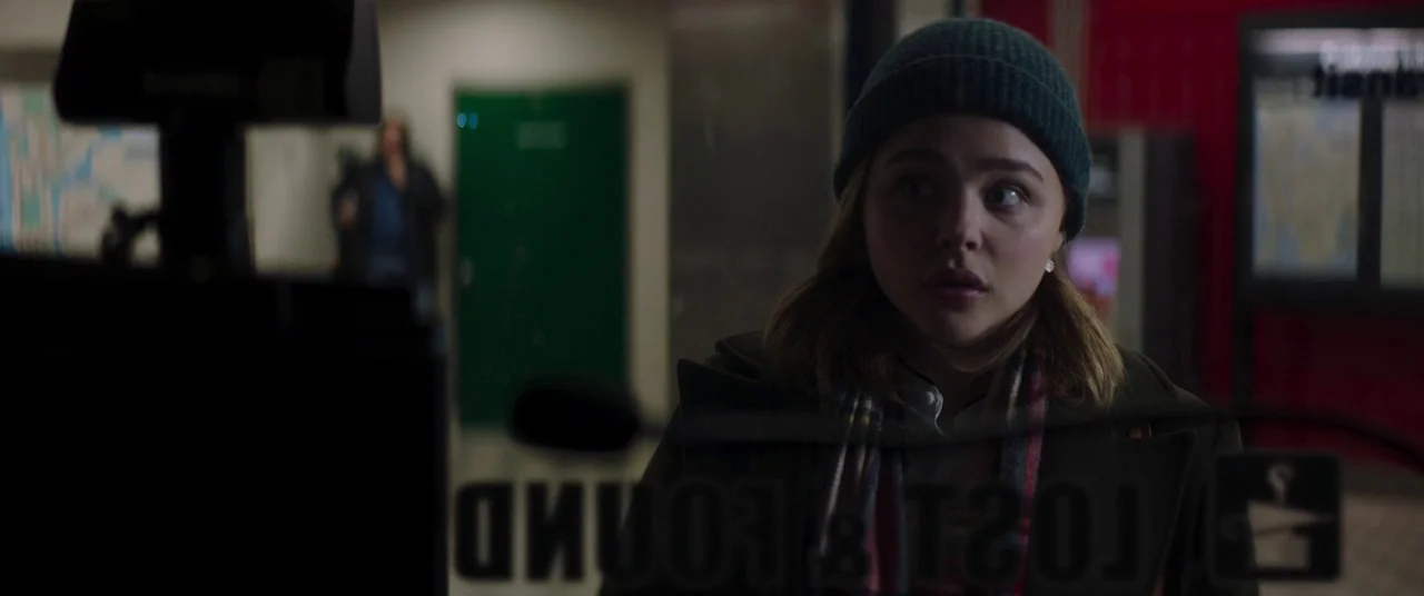 Greta movie still: medium — A dimly lit room contains a glass window with reversed text reading lost and found. A young woman in a beanie looks…