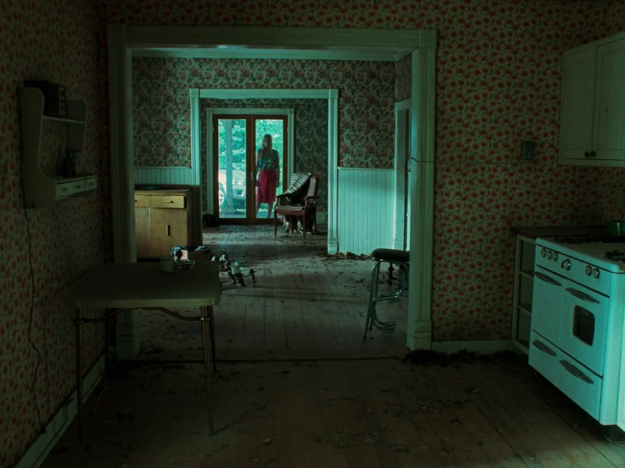 Die My Love (2025) movie still: wide — A neglected home interior features floral wallpaper and dust-covered wooden floors. A woman stands framed in…