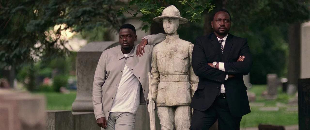 Widows (2018) movie still: medium — A quiet cemetery with scattered granite headstones and mature green trees in the background. Two men stand…