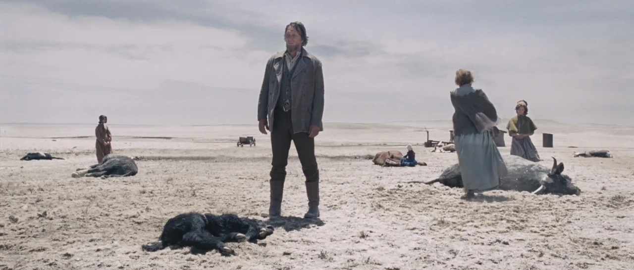 The Homesman (2014) movie still: wide — A vast, arid, light-colored landscape under a cloudy sky with scattered dead animals. Several people in…
