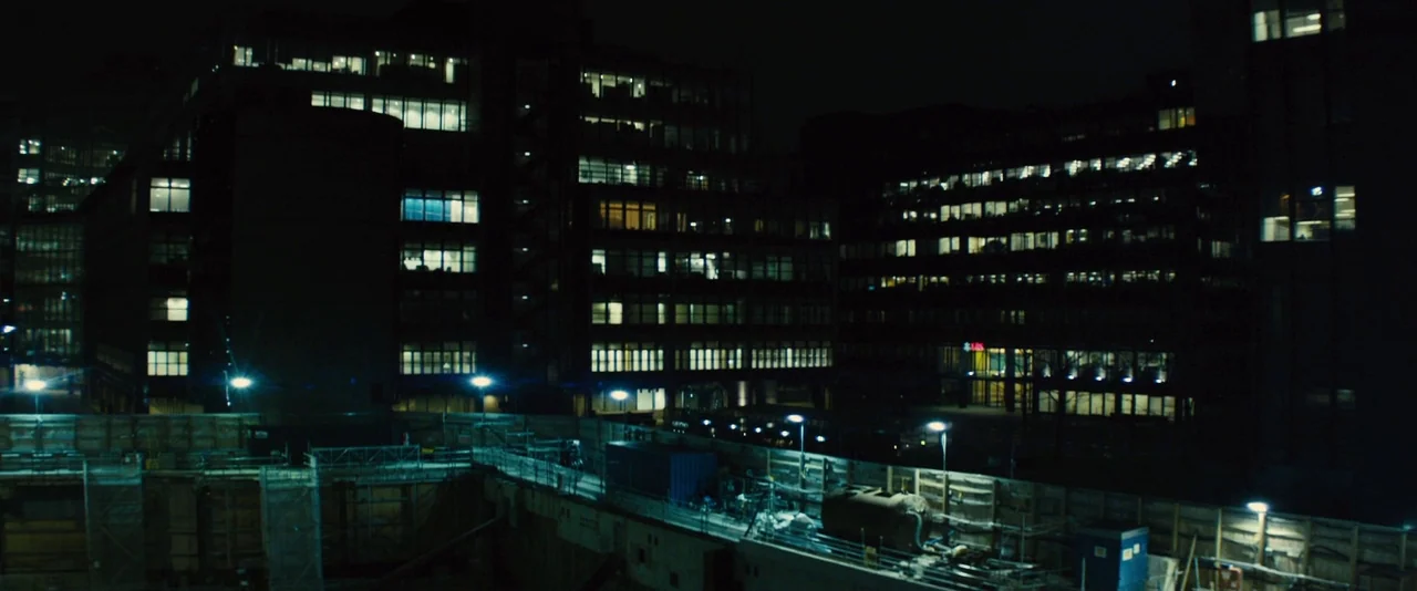 Locke (2013) movie still: establishing — A dark urban construction site sits beneath tall, multi-story office buildings at night. Rows of…