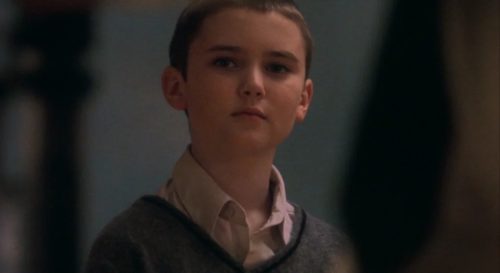 Birth (2004) movie still: closeup — A dimly lit indoor space with blurred background walls of blue-gray tone. A young boy in a v-neck sweater turns…