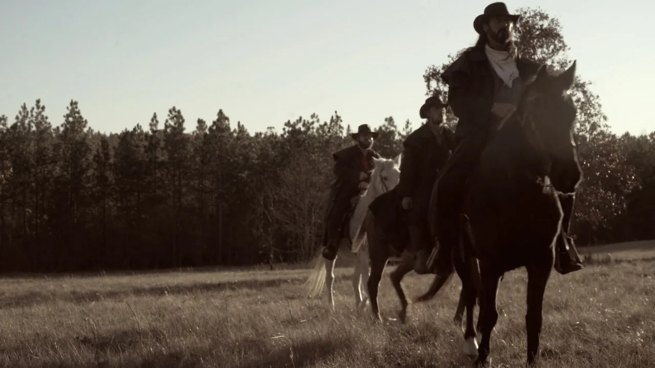 The Dalton Gang (2020) movie still: wide — Three mounted men travel through a grassy field bordered by a dense pine forest. The riders wear…