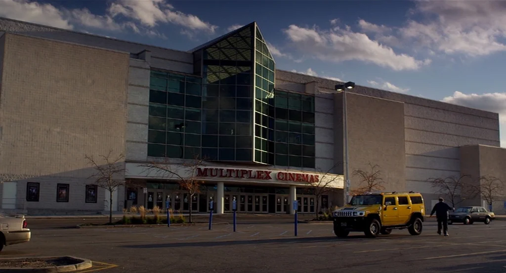 Dark Horse (2011) movie still: establishing — A sprawling, multi-story multiplex cinema building stands at the edge of a parking lot. A bright…