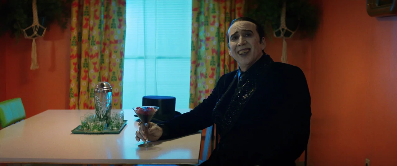 Renfield (2023) movie still: medium — A dining room with bright orange walls and patterned curtains flanking a bright window. A man sits at a white…