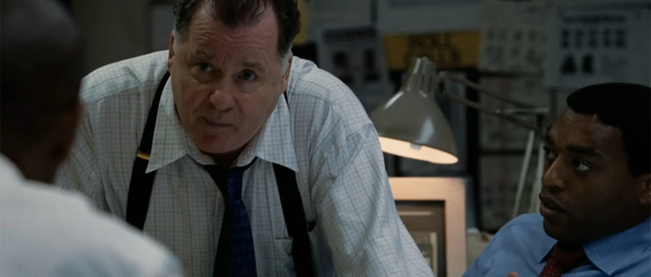 Inside Man (2006) movie still: medium — A cluttered office workspace contains papers, desk lamps, and documents pinned to walls. Two men sit in a…