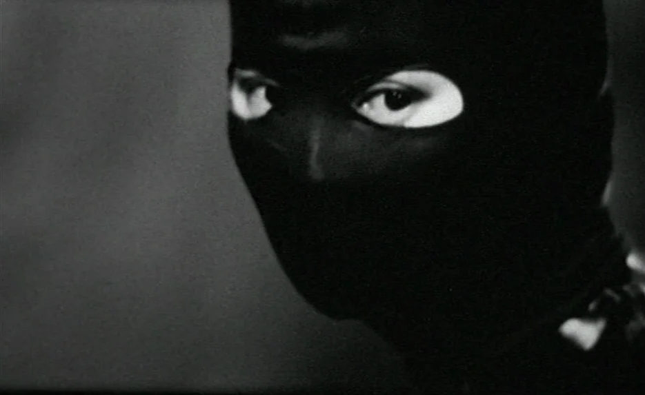 Irma Vep (1996) movie still: closeup — A dark, undifferentiated background features a single person wearing a close-fitting black mask. The person…