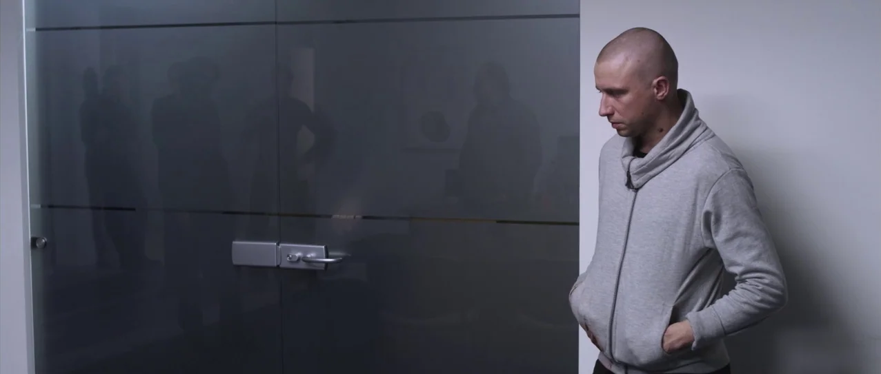 Vanishing Waves (2012) movie still: medium — A man stands in an office corridor beside a large dark glass double door. The glass reflects the faint…