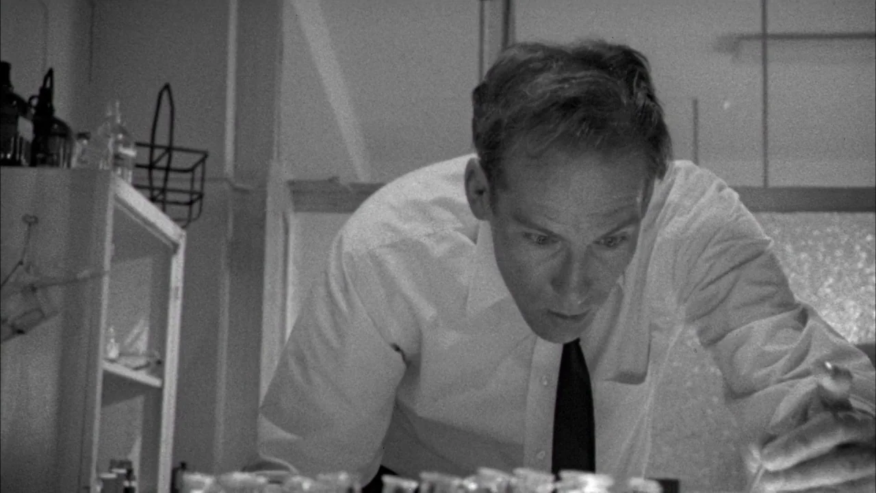 Poison movie still: medium — A scientific workspace contains shelves filled with glass beakers and chemical bottles. A man in a dress shirt leans…