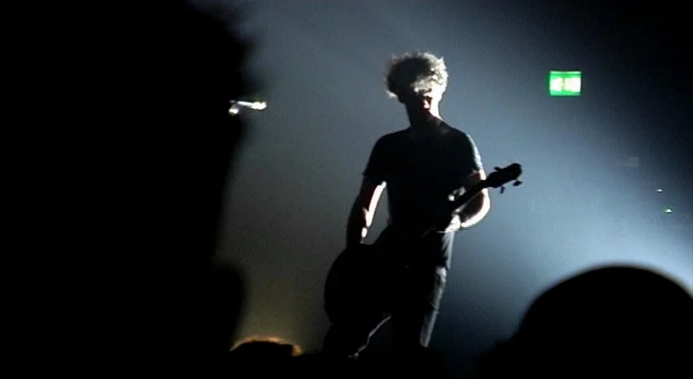 9 Songs (2004) movie still: fullbody — A dimly lit room features a performer on a stage. The subject plays a guitar while standing in front of a…
