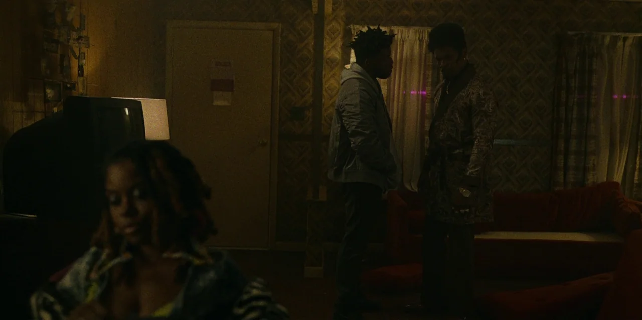 They Cloned Tyrone (2023) movie still: medium — A dimly lit room with 1970s patterned wallpaper and a prominent red couch. Two men stand in the…