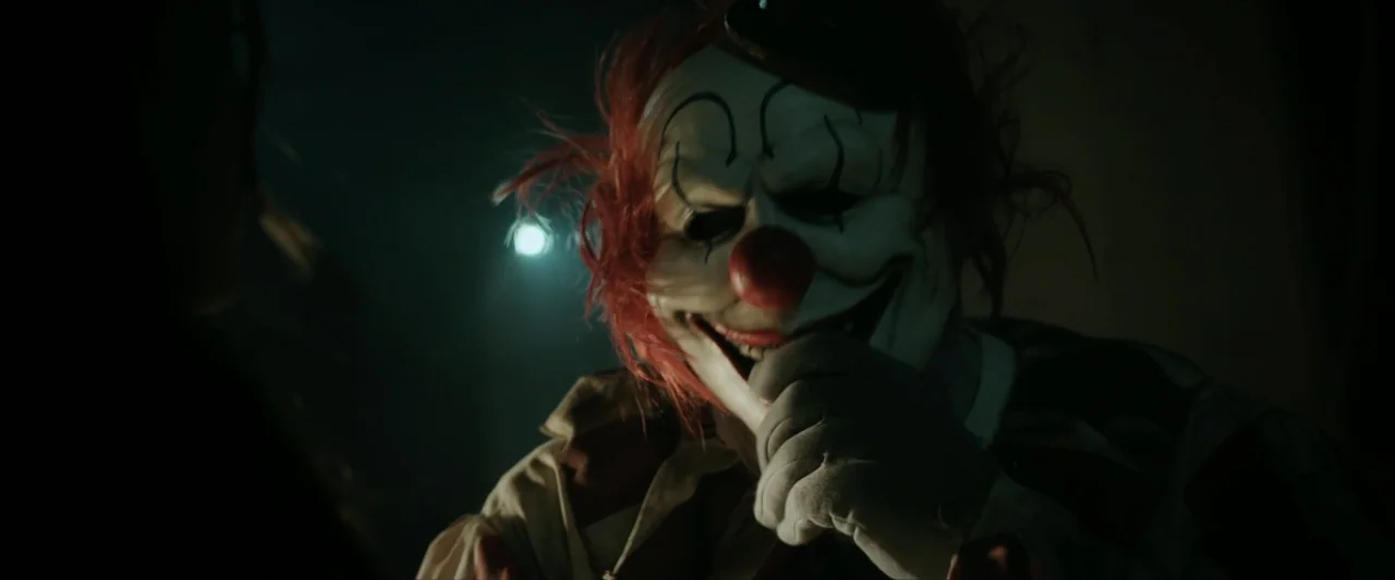 Clown in a Cornfield (2025) movie still: closeup — A dark, dimly lit indoor space frames a character wearing a distressed clown mask. The masked…
