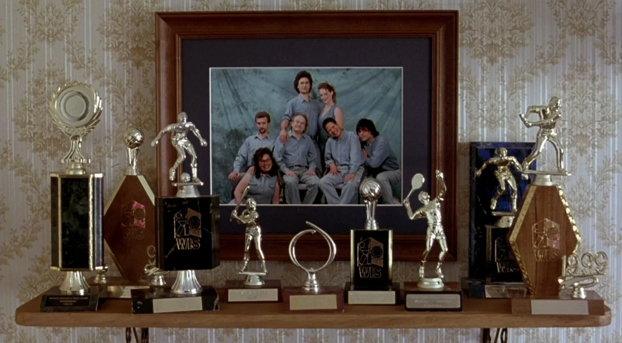 Eagle Vs. Shark movie still: medium — A wooden shelf holds a row of various sport trophies against damask-patterned wallpaper. A framed group…