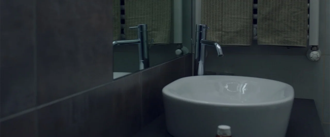 About Us (2017) movie still: medium — A modern bathroom features dark grey tile walls and a white ceramic vessel sink. A clean countertop supports a…