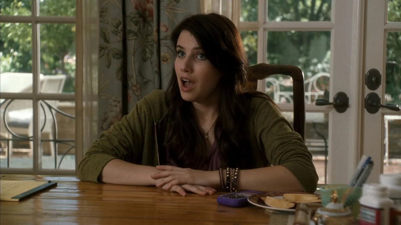 F Valentine's Day (2026) movie still: medium — A sunny dining room with white French doors and floral curtains. A young woman sits at a wooden table…