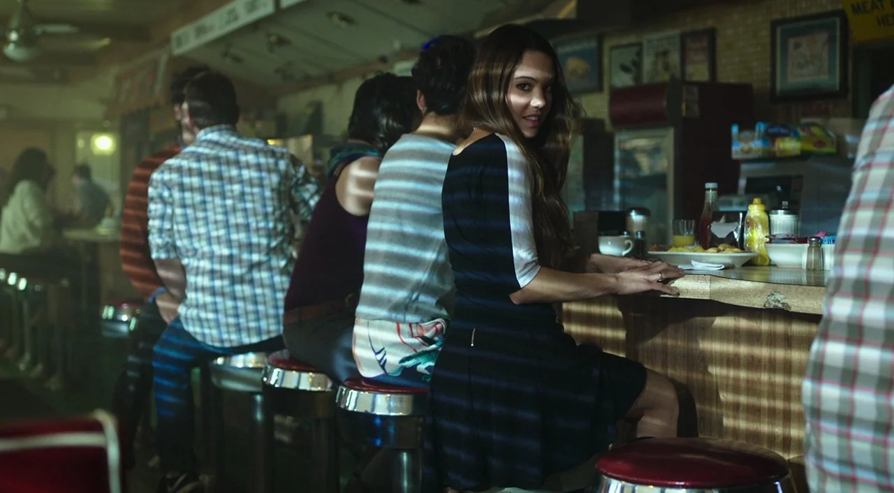 Urge (2016) movie still: medium — A classic diner interior with multiple patrons sitting on red stools at the counter. A young woman in a striped…