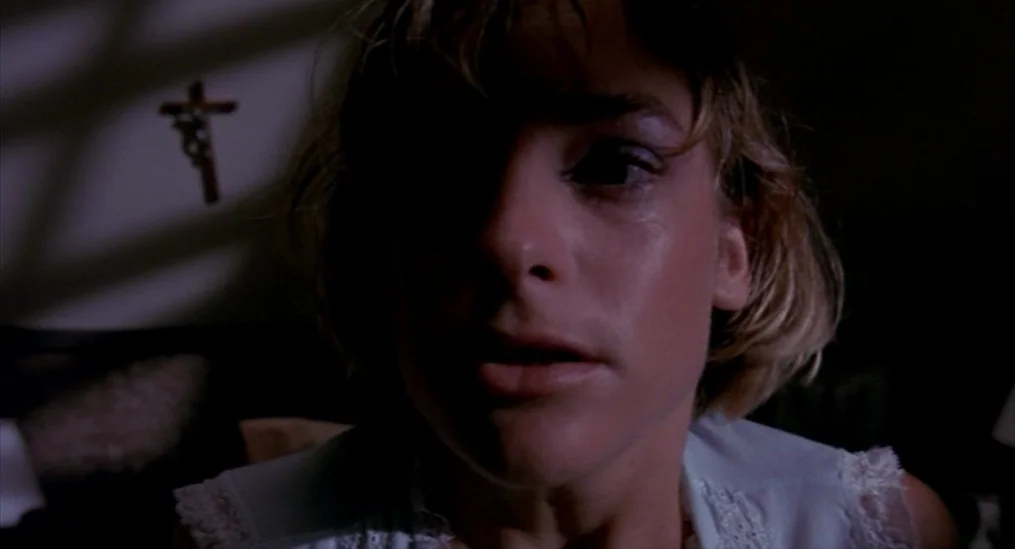 A Nightmare On Elm Street (1984) movie still: closeup — A dark, dimly lit room features a wall with a wooden crucifix mounted above. A young woman…
