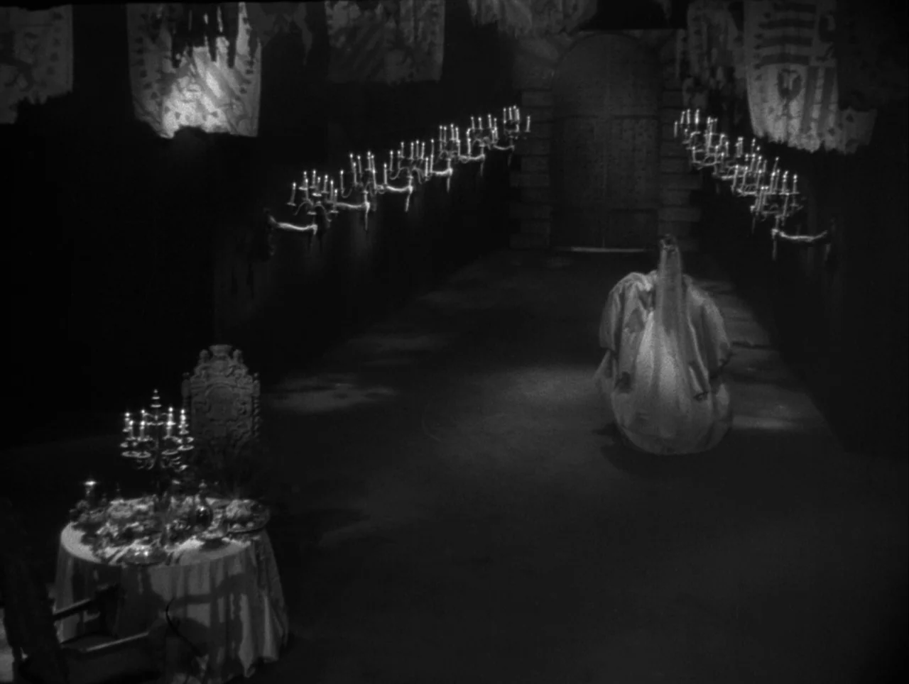 La Belle et la Bête (1946) movie still: wide — A dark stone hall features rows of wall-mounted candles and heraldic banners hanging above. A…