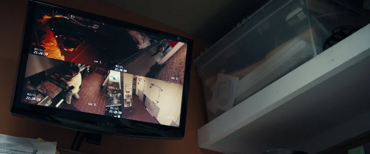 Compliance (2012) movie still: medium — A dark corner room holds a wall-mounted security monitor displaying four distinct video feeds. One screen…