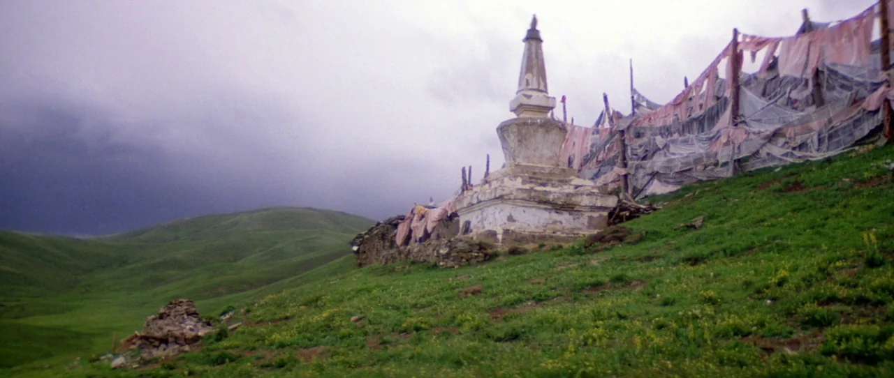 Horse Thief movie still: wide — A stone stupa stands on a green, grassy slope under a dark, cloudy sky. Faded prayer flags are draped across a fence…