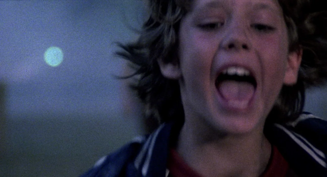 City of the Living Dead (1980) movie still: closeup — A dark, blurred outdoor space features a glowing moon in the distance. A young boy runs past…