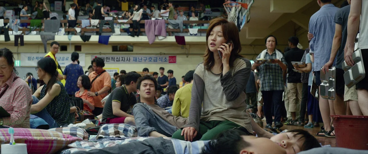 Parasite (2019) movie still: medium — A crowded temporary shelter occupies a large indoor gymnasium filled with people sleeping on mats. A woman…