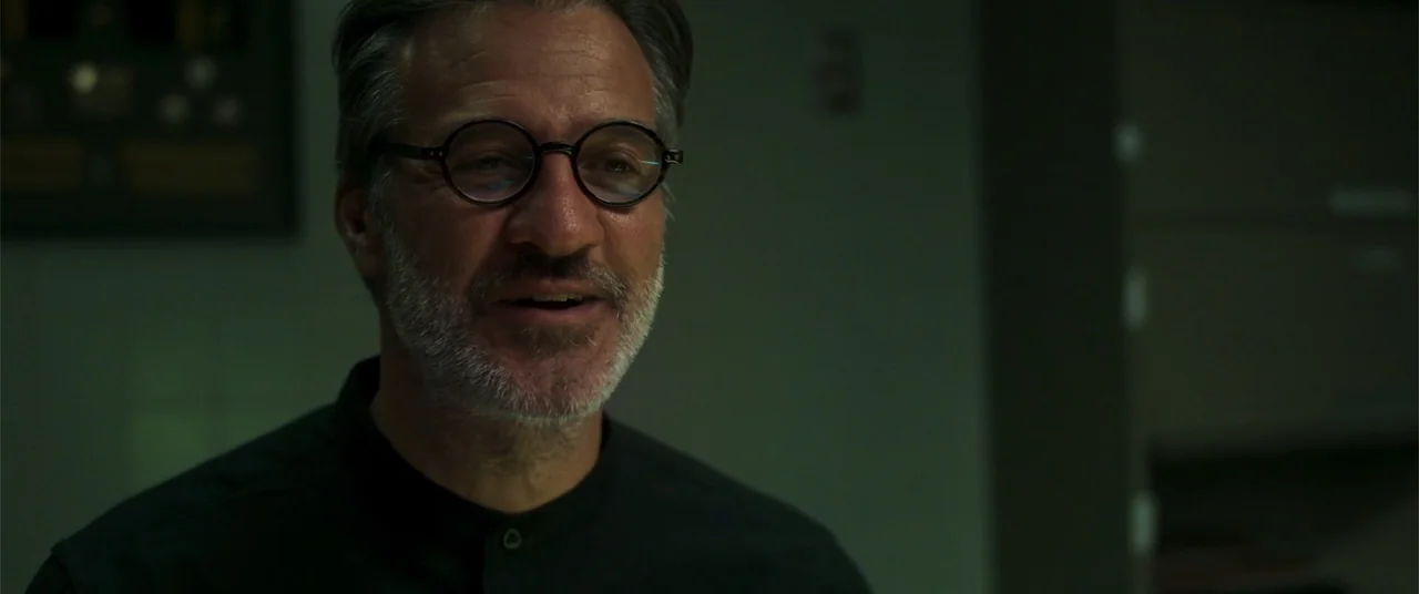 Phobias (2021) movie still: closeup — A dimly lit professional space features dark walls and minimal environmental detail. A middle-aged man with a…