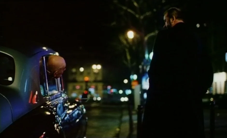 Simon, The Magician (1999) movie still: medium — A dark urban street at night illuminated by distant, blurred city lights. A man inside a car leans…