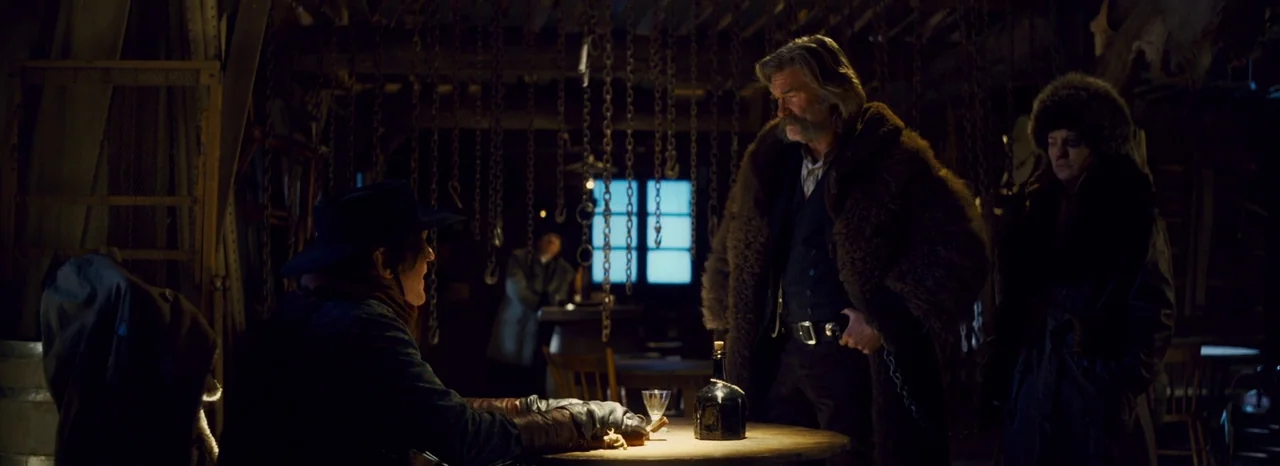 The Hateful Eight (2015) movie still: wide — A dimly lit, rustic interior features hanging chains and wooden tables in the background. A man in a…