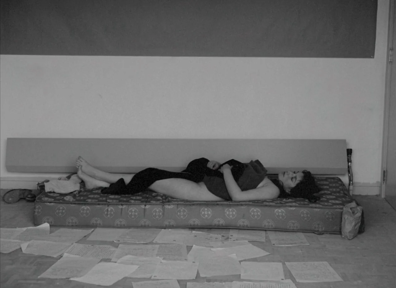 Je Tu Il Elle (1974) movie still: fullbody — An unfurnished room has white walls and a bare floor covered in scattered papers. A woman sleeps on a…