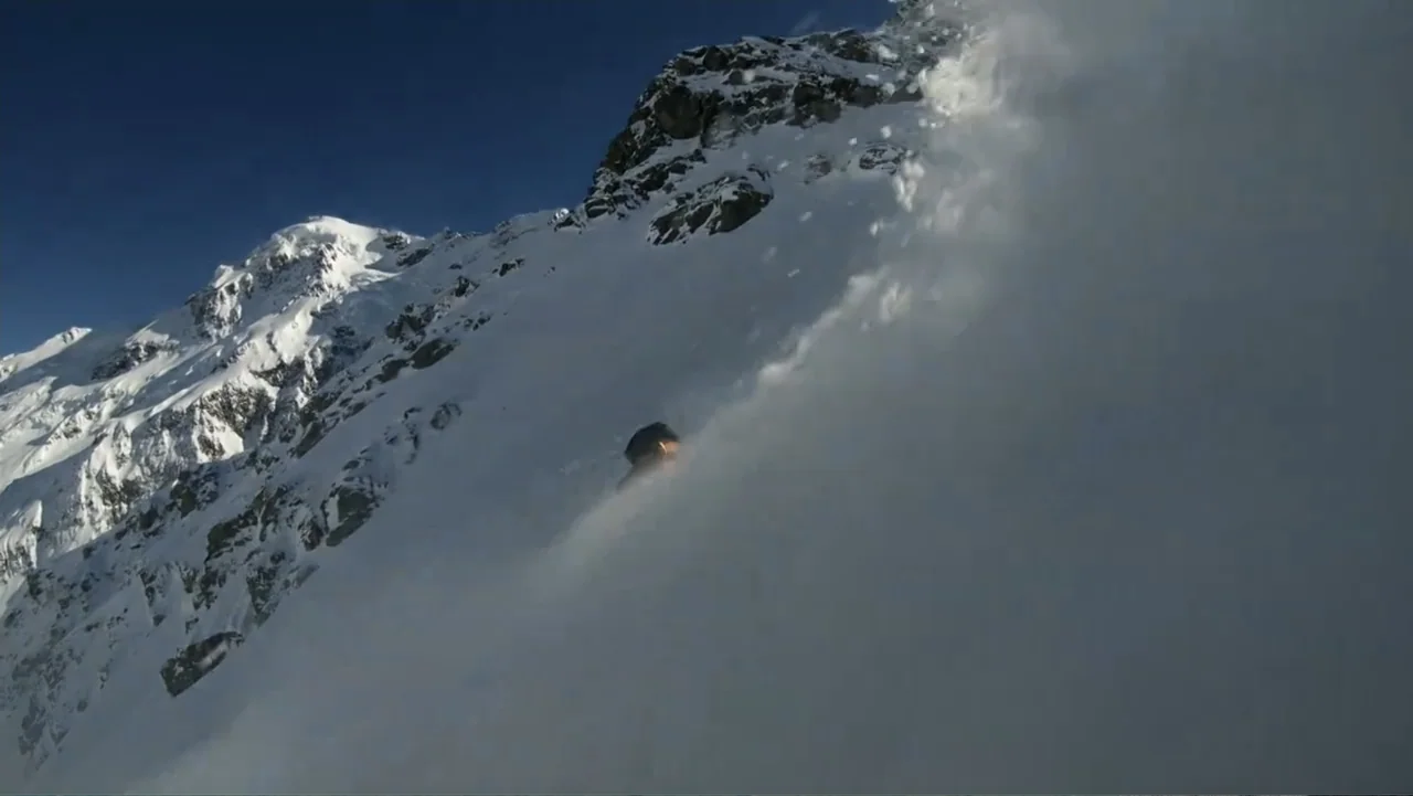 Dear Rider: The Jake Burton Story (2021) movie still: wide — A steep snow-covered mountain slope rises under a clear deep blue sky. A solitary skier…