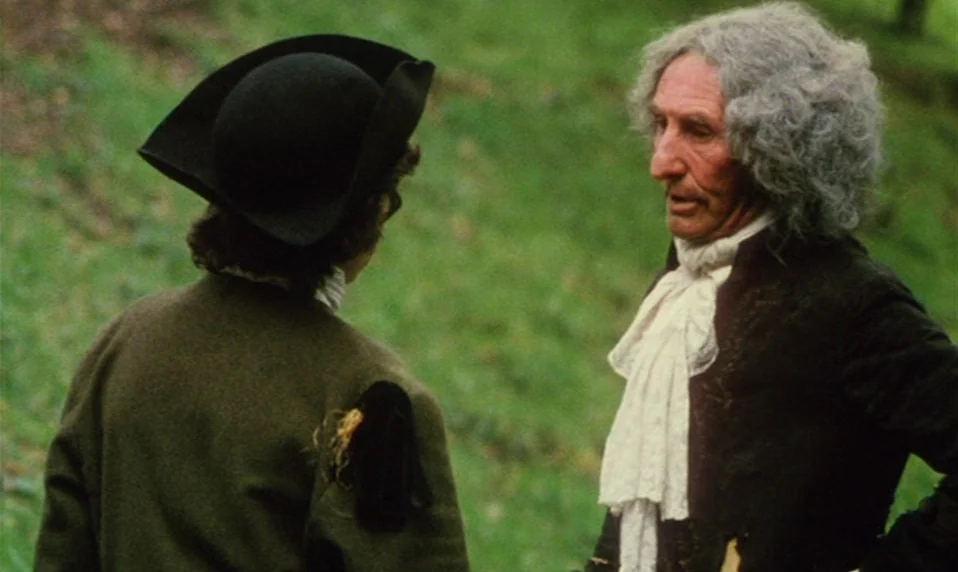 Black Jack (1979) movie still: medium — Two men dressed in 18th-century attire converse in a verdant, grassy outdoor field. One man wears a period…