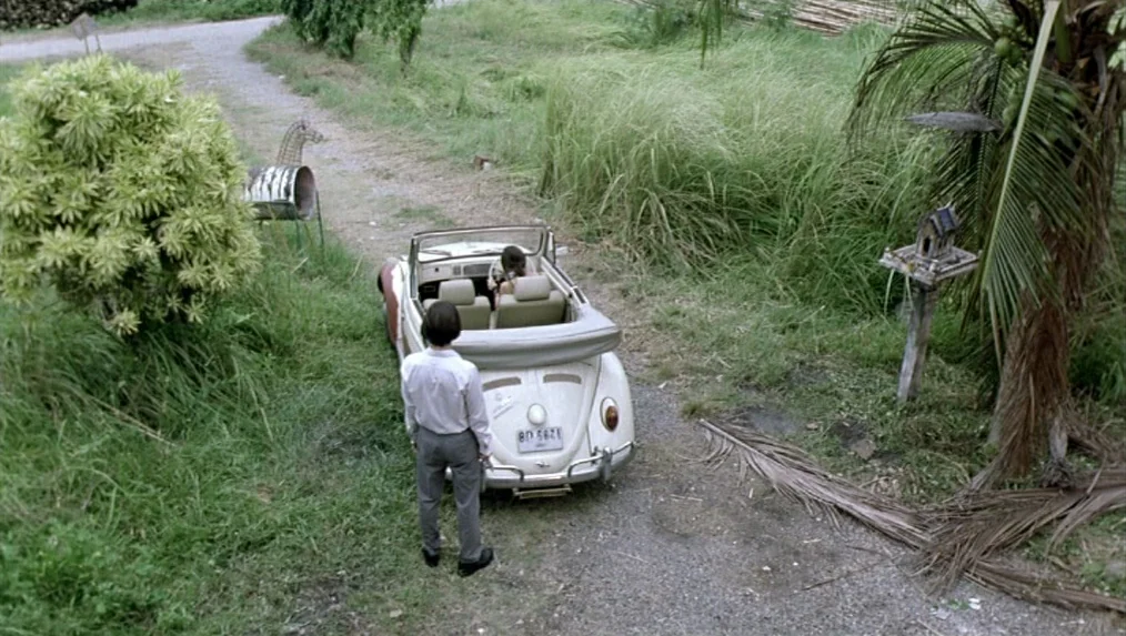 Last Life in the Universe (2003) movie still: wide — A rural dirt path winds through lush tall grasses with a white convertible parked. The man…