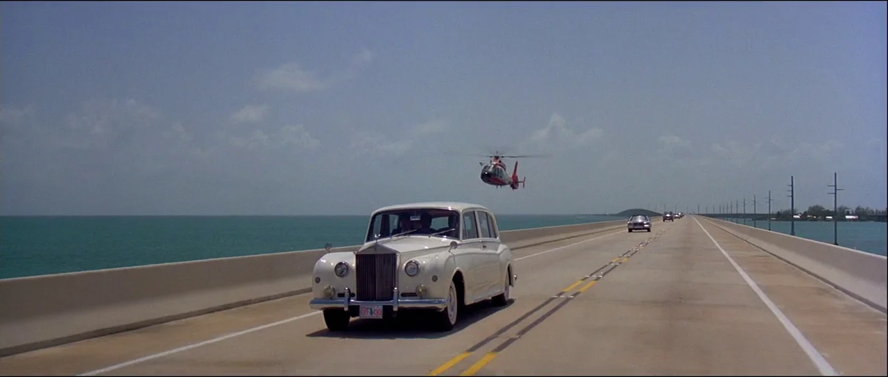 Licence to Kill (1989) movie still: wide — A long concrete bridge stretches across turquoise open water under a clear sky. A vintage white Rolls…
