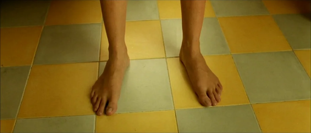 Amelie (2001) movie still: closeup — A floor is covered in alternating yellow and sage green square tiles. Bare human feet stand still on the tiled…