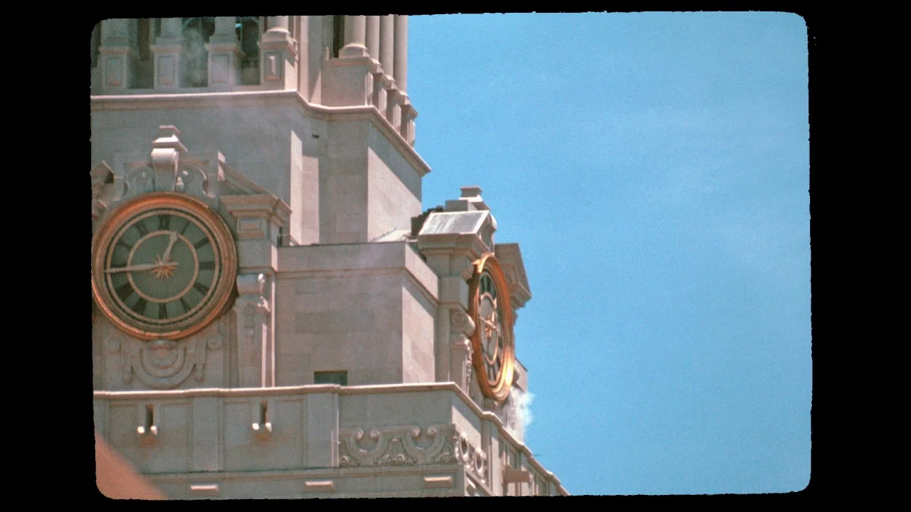 Tower (2016) movie still: establishing — The stone exterior of a neoclassical clock tower features two prominent circular clock faces. Sunlight…