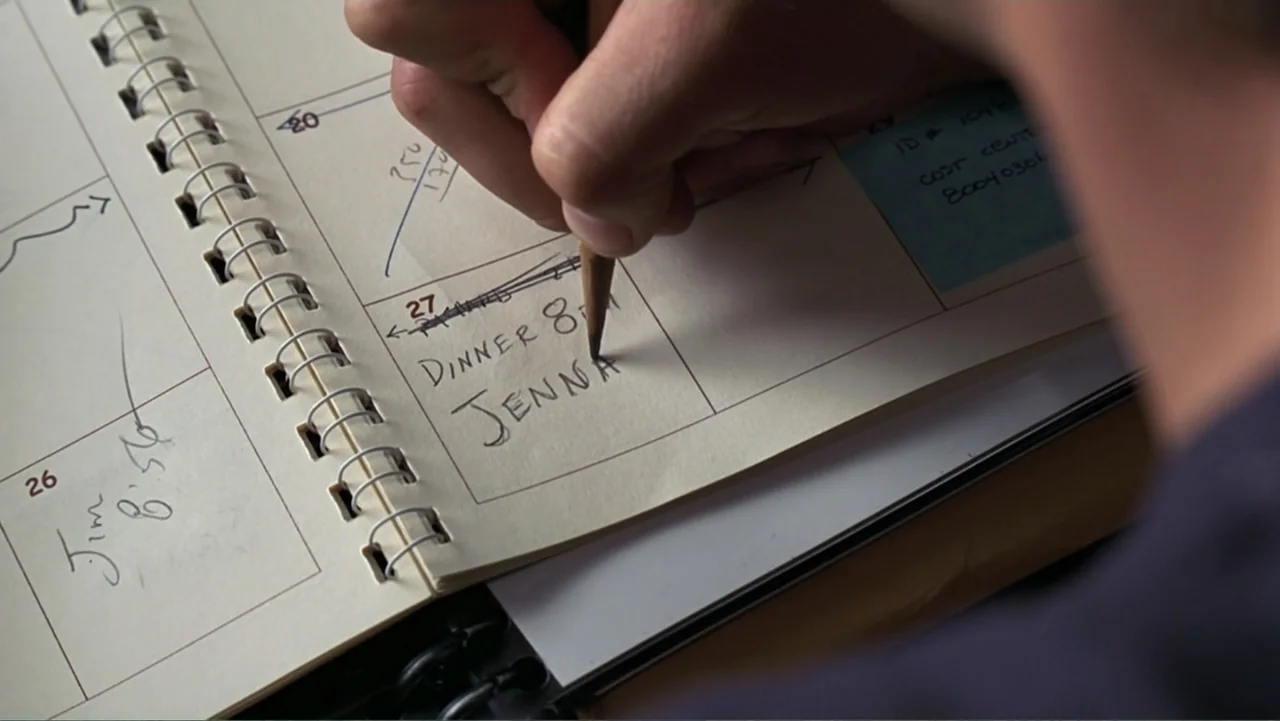 13 Going on 30 (2004) movie still: closeup — An open spiral-bound appointment book rests on a flat wooden desk surface. A hand holds a wooden pencil…