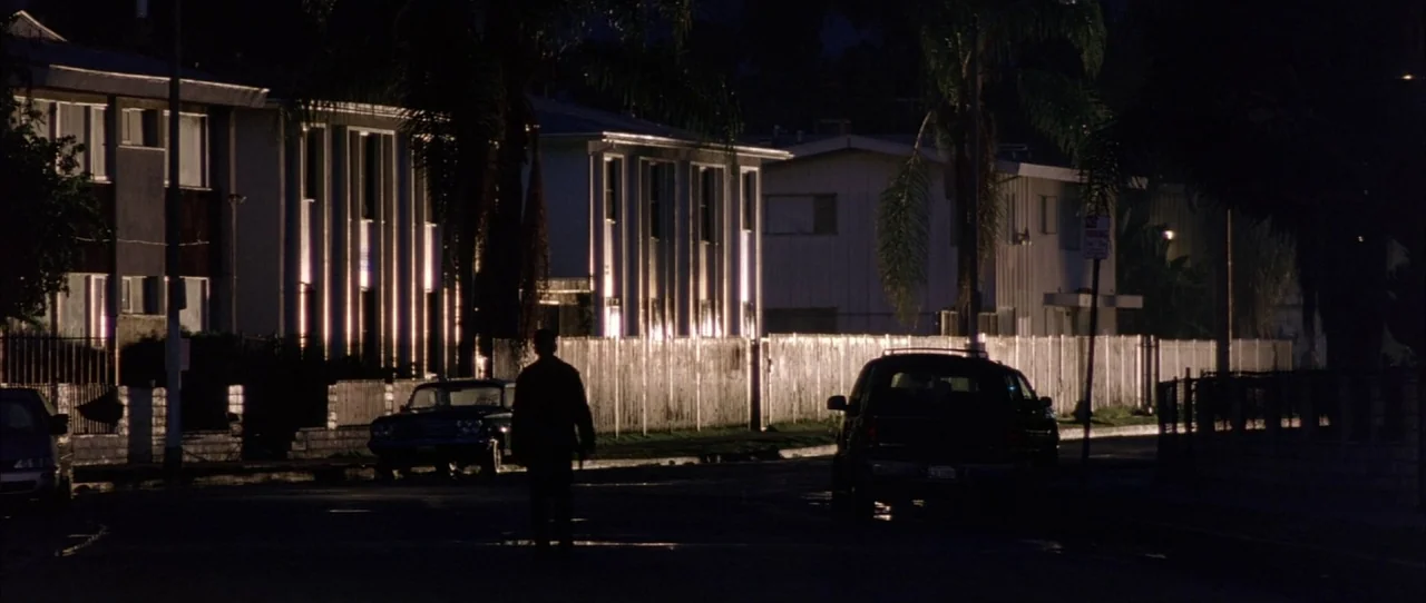 Training Day (2001) movie still: wide — A dimly lit street lined with modern apartment complexes and parked cars at night. A lone man stands facing…