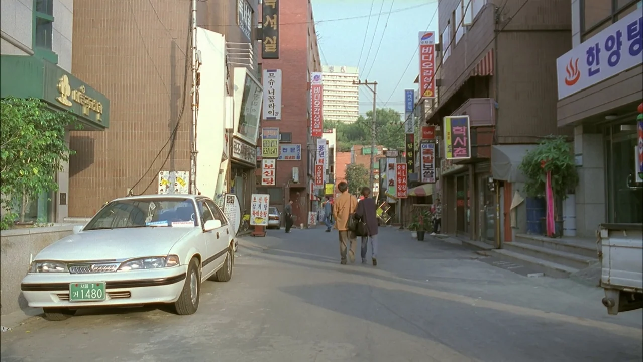 The Day a Pig Fell Into the Well (1996) movie still: wide — A narrow residential street in South Korea is flanked by multistory brick buildings. A…