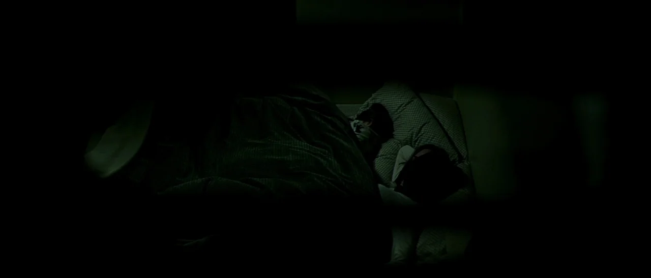 Dark Water (2005) movie still: medium — A dark bedroom features two people sleeping in a bed under a patterned blanket. The figures are partially…