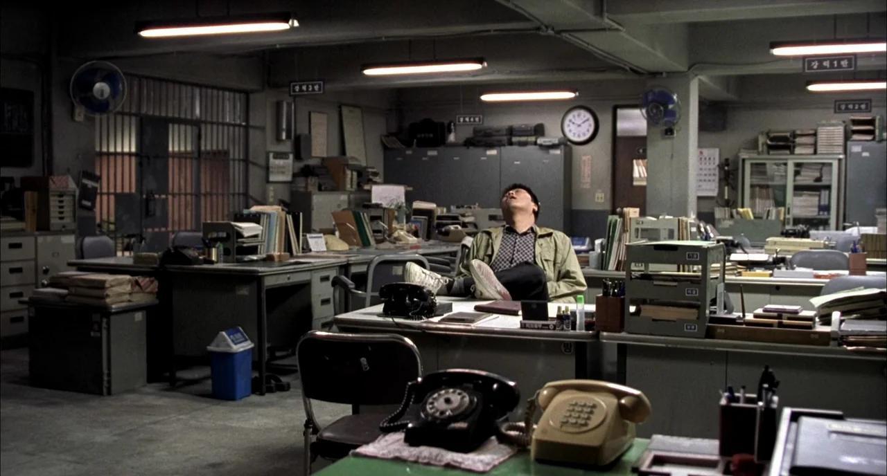 Memories of Murder (2003) movie still: wide — A cluttered government office features rows of gray metal desks stacked with paper files. A man sleeps…