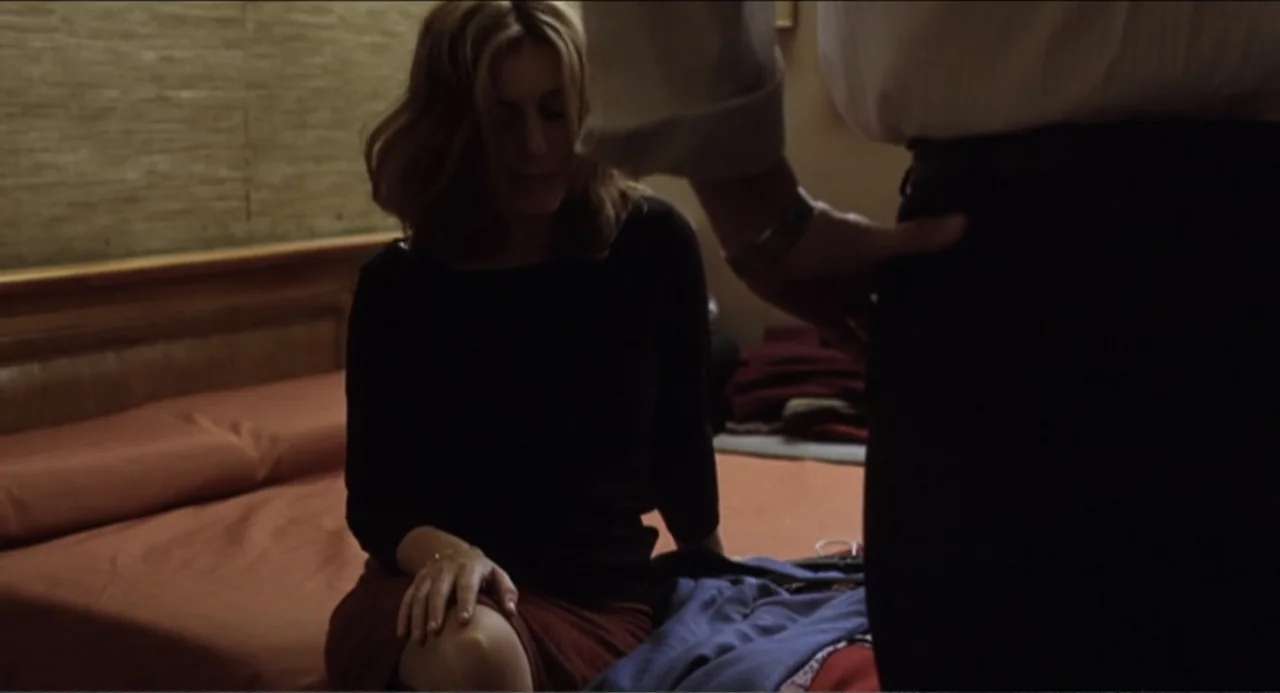 The Holy Girl (2004) movie still: medium — A furnished room contains a bed with a plain headboard and textured walls. A woman sits on the edge of…