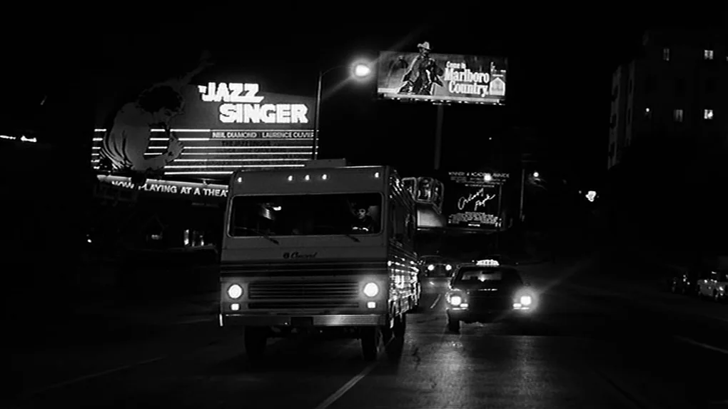 The State of Things (1982) movie still: wide — An urban street at night is illuminated by car headlights and glowing advertising billboards. A…