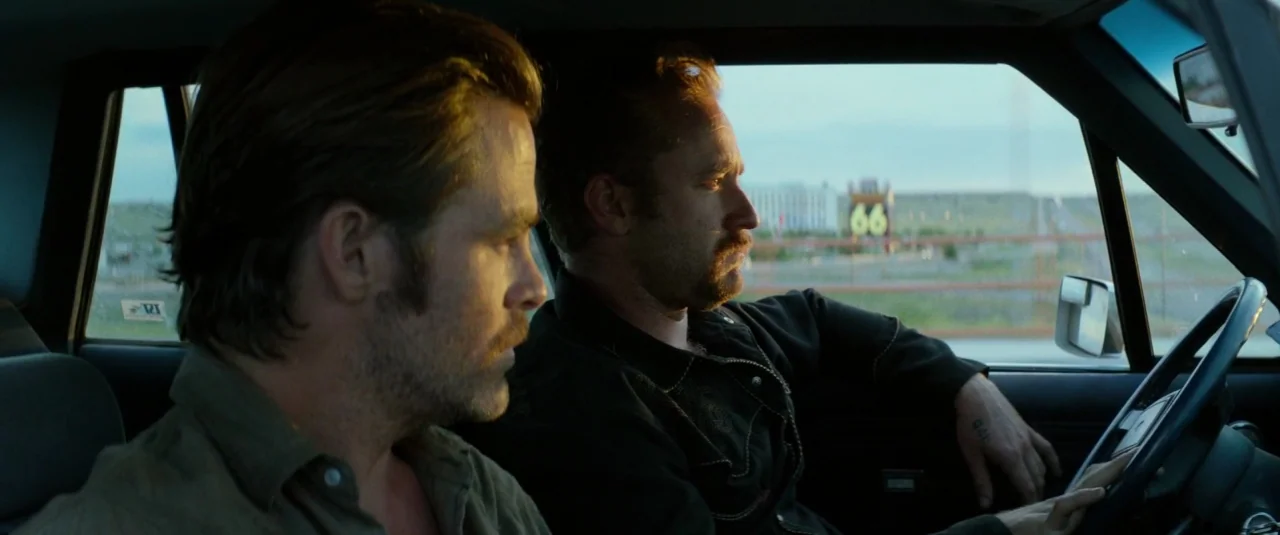 Hell or High Water (2016) movie still: medium — Two men sit inside a car cabin during the golden hour of the day. A blurred highway sign marked 66…