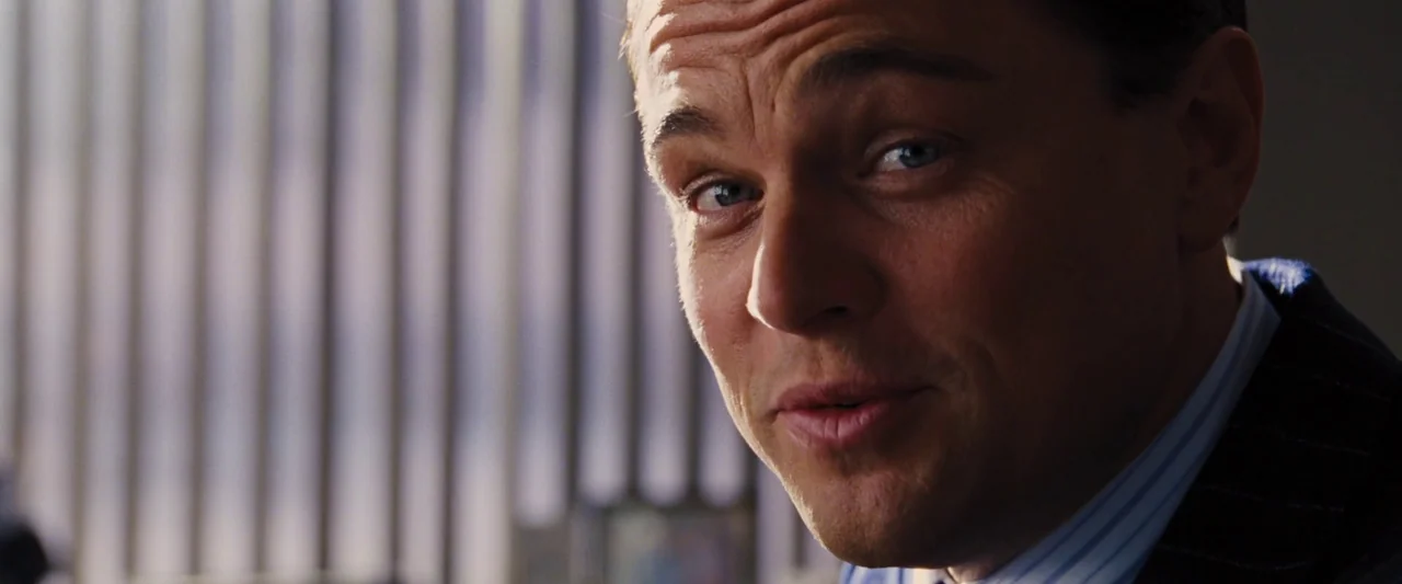 The Wolf of Wall Street (2013) movie still: closeup — An office interior features vertical blinds covering a bright window in the background. A man…