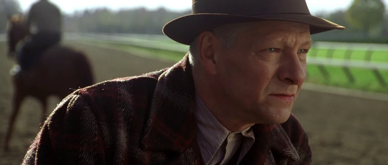 Seabiscuit (2003) movie still: medium — A dirt horse racing track with a green infield and a white fence. A man in a fedora watches a rider on a…