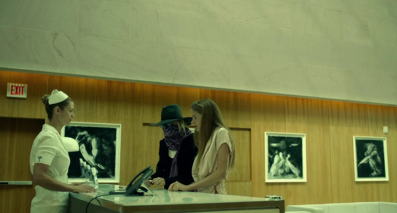 Rabid (2019) movie still: medium — A reception area with wood paneled walls decorated with framed black and white illustrations. A nurse standing…