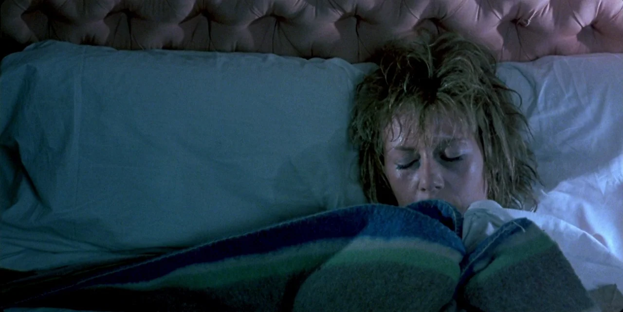 Dark Habits (1983) movie still: closeup — A person lies on a white pillow against a pink tufted headboard. A blue and green striped blanket covers…
